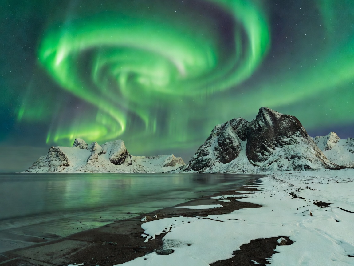 Green northern lights over a snow-dusted Lofoten beach with steep mountains rising behind the shoreline